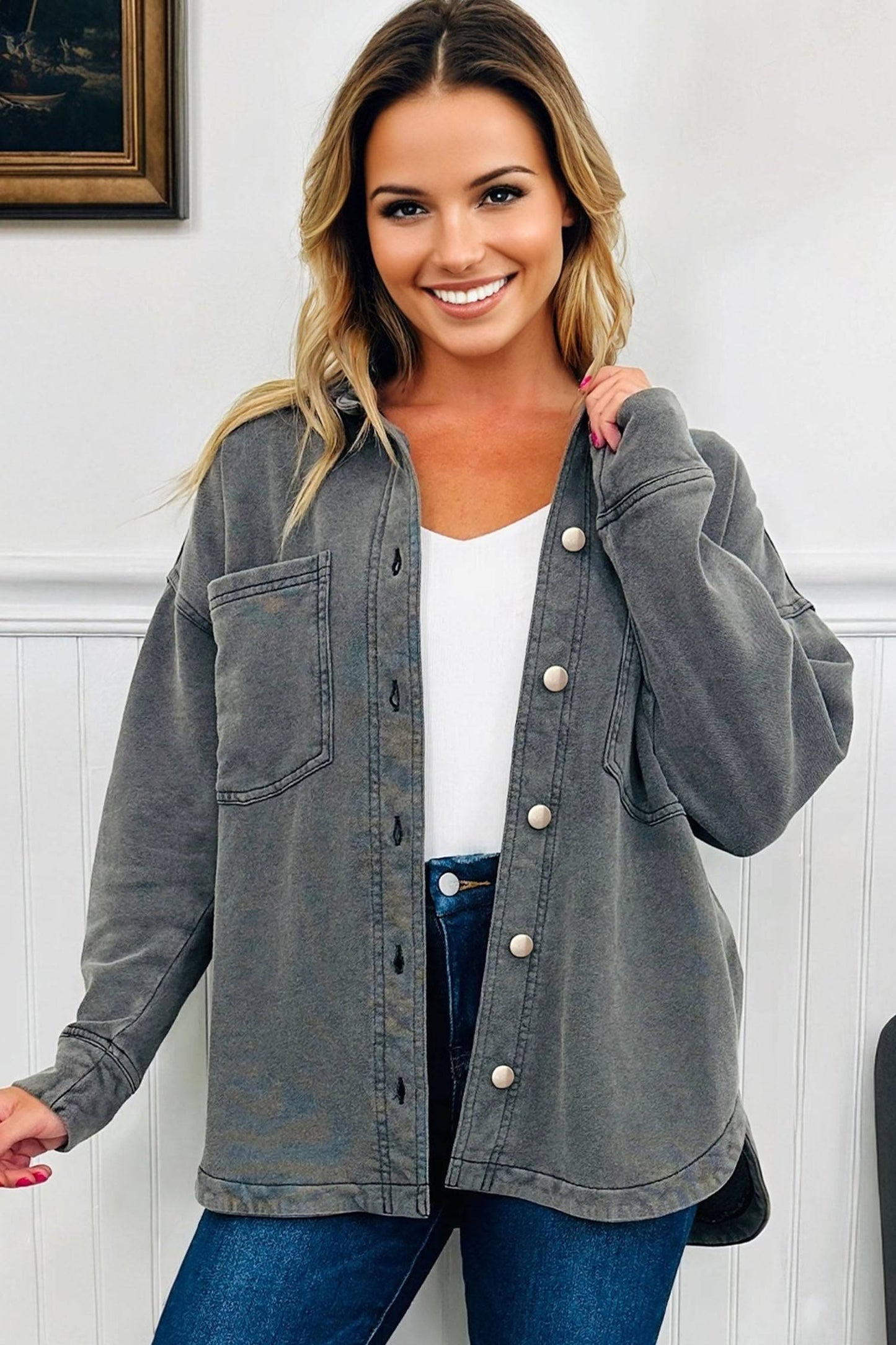 Woman wearing a gray denim jacket over a white top and blue jeans, standing against a white wall.