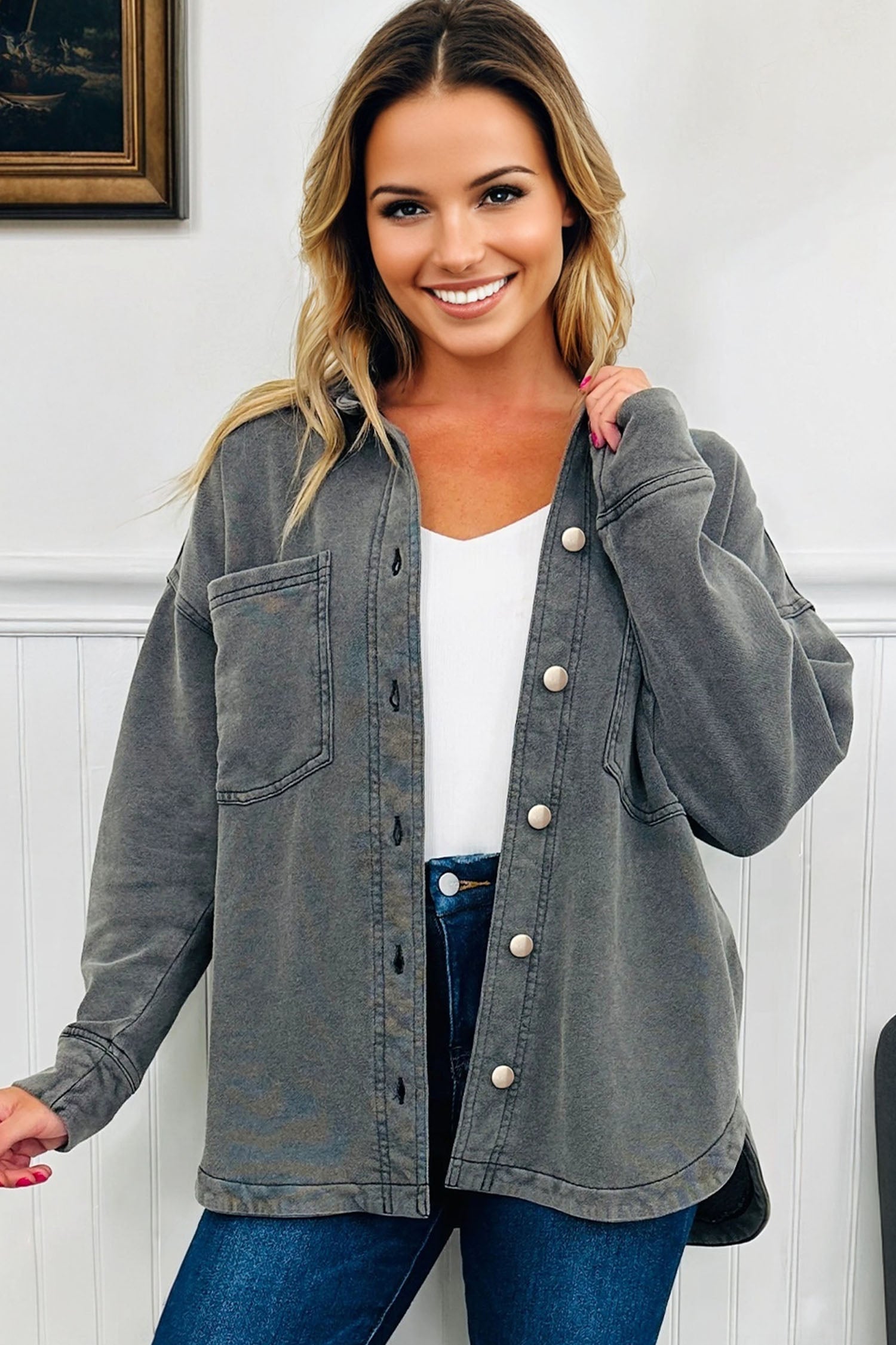 Woman wearing a gray denim jacket over a white top and blue jeans, standing against a white wall.