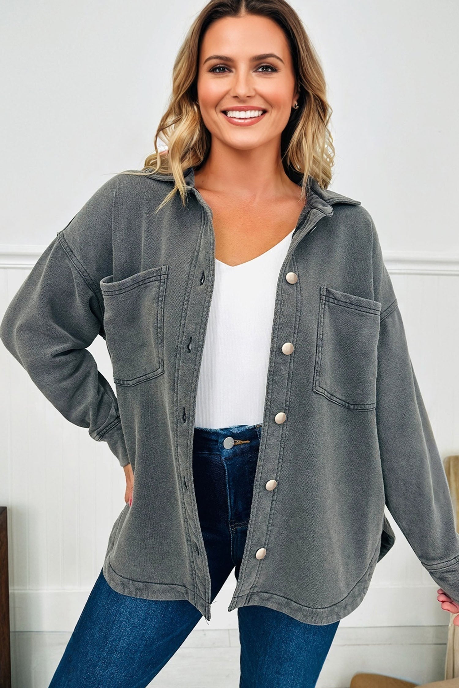 Woman wearing a gray button-up shirt over a white top and blue jeans, standing against a white wall.