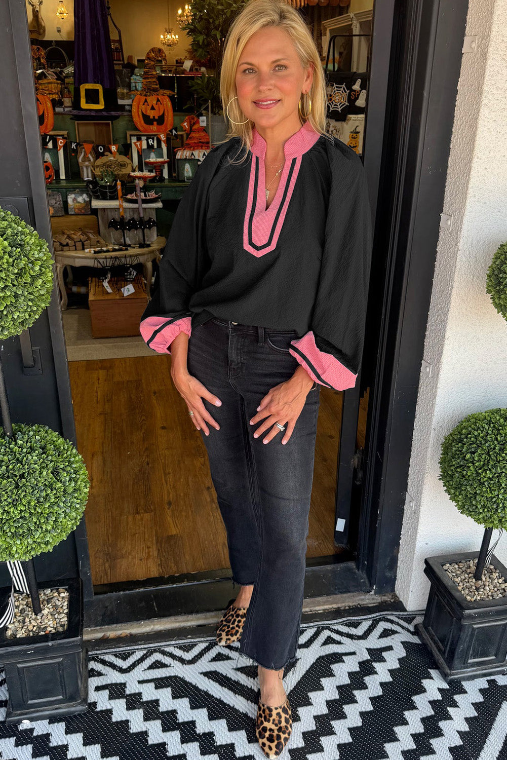 Woman standing outside a store with Halloween decorations, wearing a black top with pink accents and dark jeans.