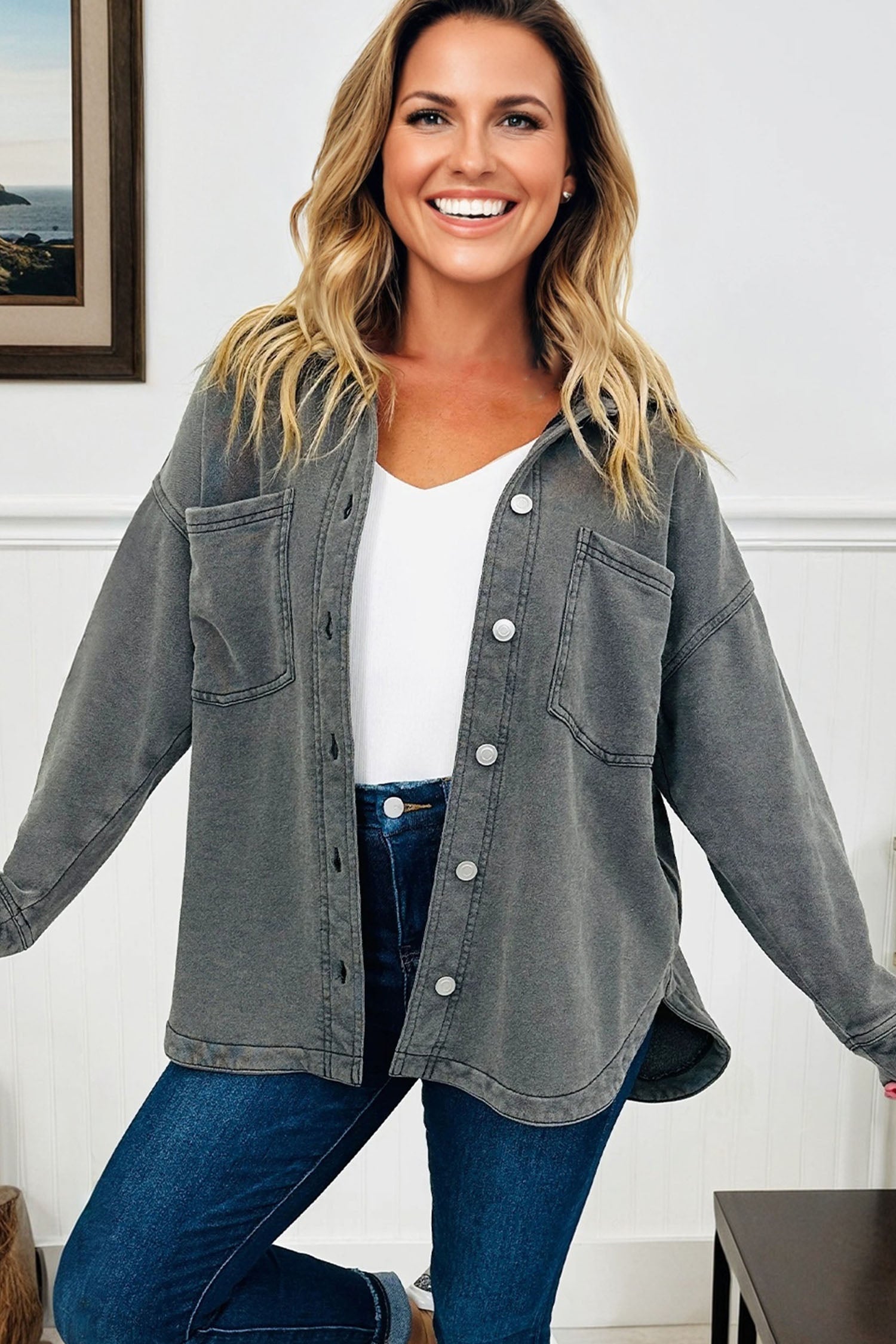 Woman wearing a gray button-up shirt over a white top and blue jeans, standing indoors.