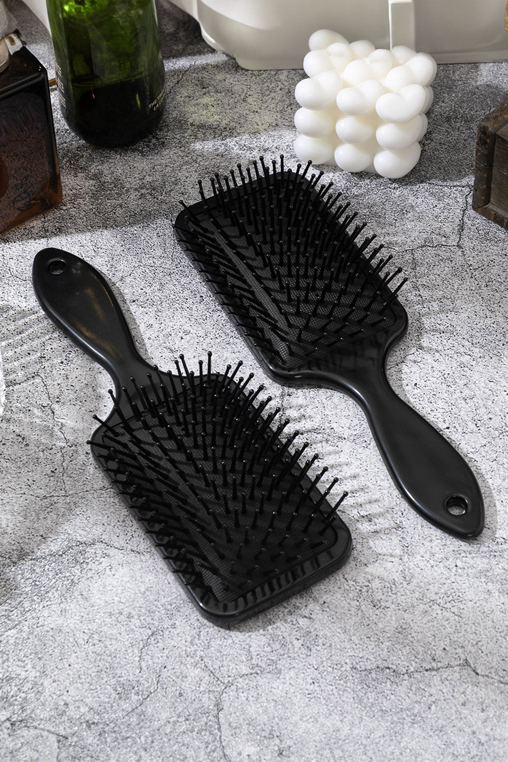 Two black hairbrushes on a textured surface with a bottle and decorative items in the background.