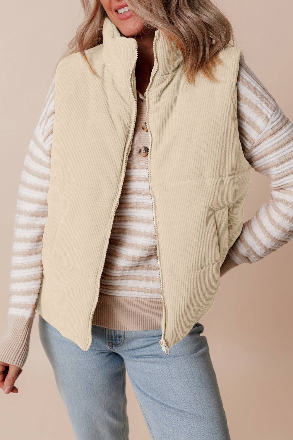 Woman wearing a beige puffer vest over a striped sweater with a beige background