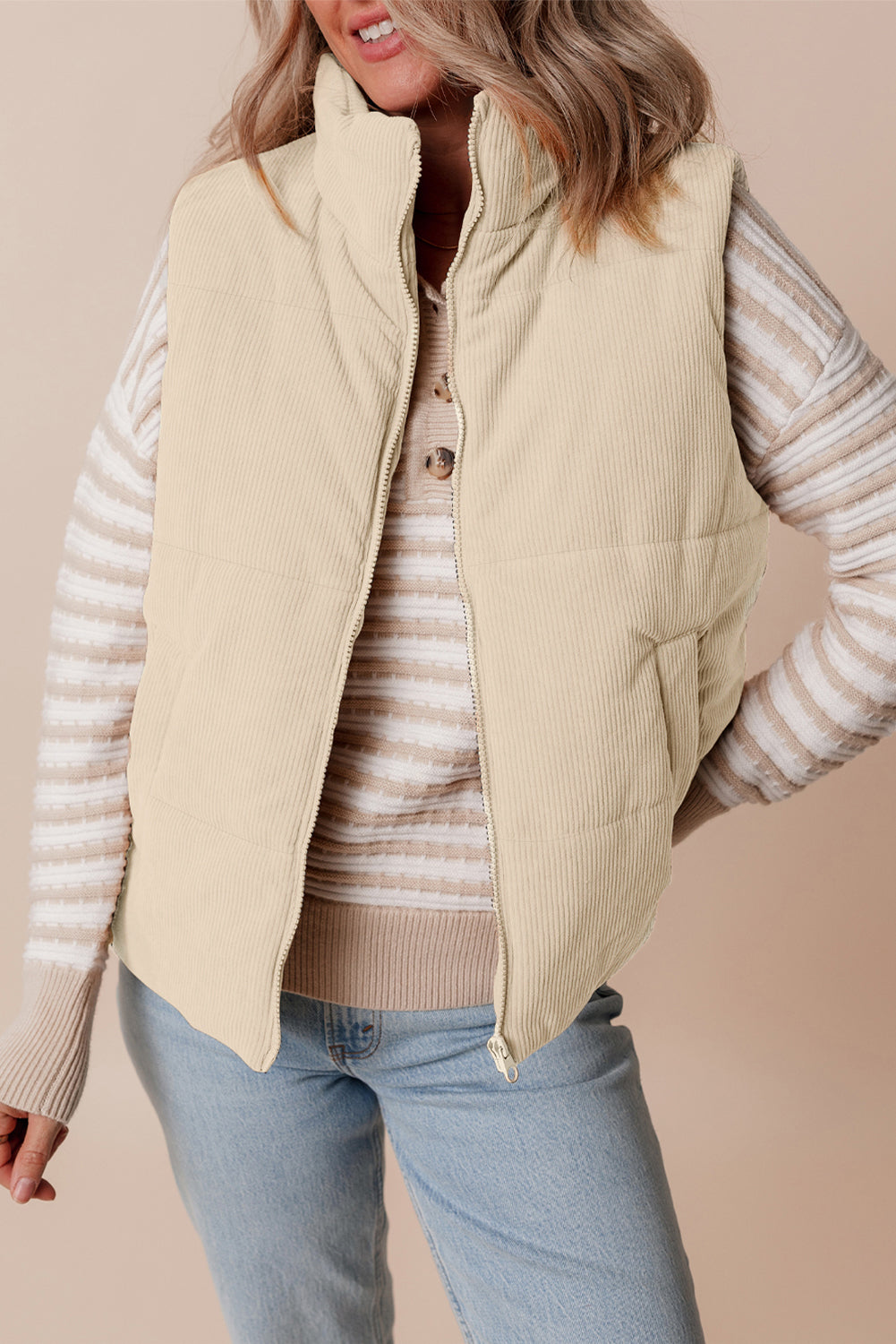 Woman wearing a beige puffer vest over a striped sweater with a beige background