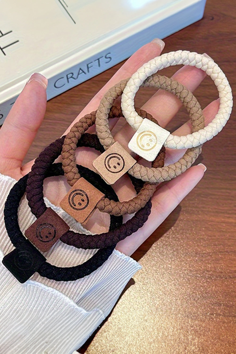 Hand holding three braided hair ties with wooden buckles on a wooden surface.