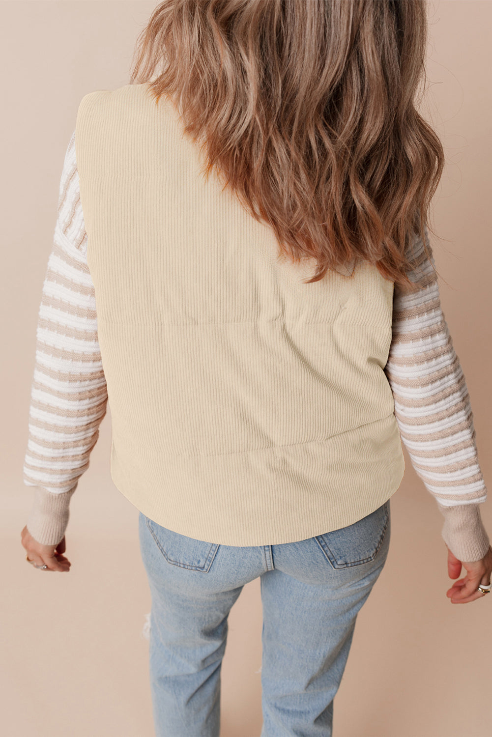 Person wearing a beige vest over a striped shirt with light blue jeans on a beige background