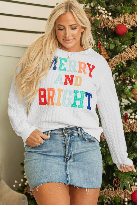 Woman wearing a white sweater with colorful text in front of a decorated Christmas tree.