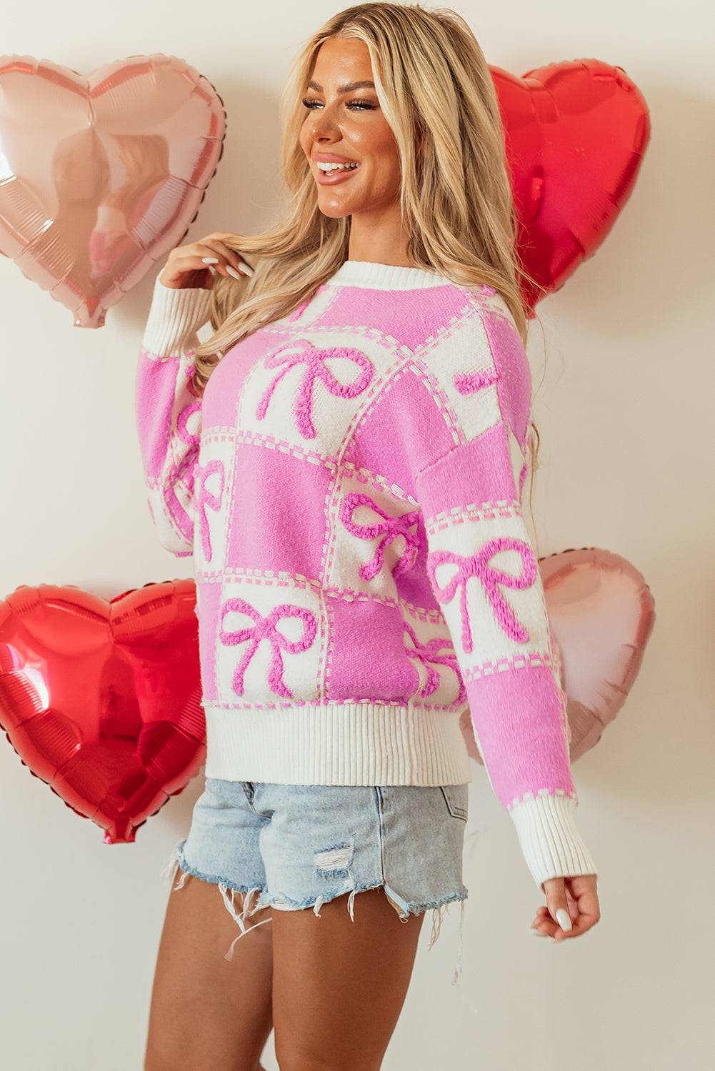Woman wearing a pink and white sweater with heart designs, standing against a light background with heart-shaped balloons.
