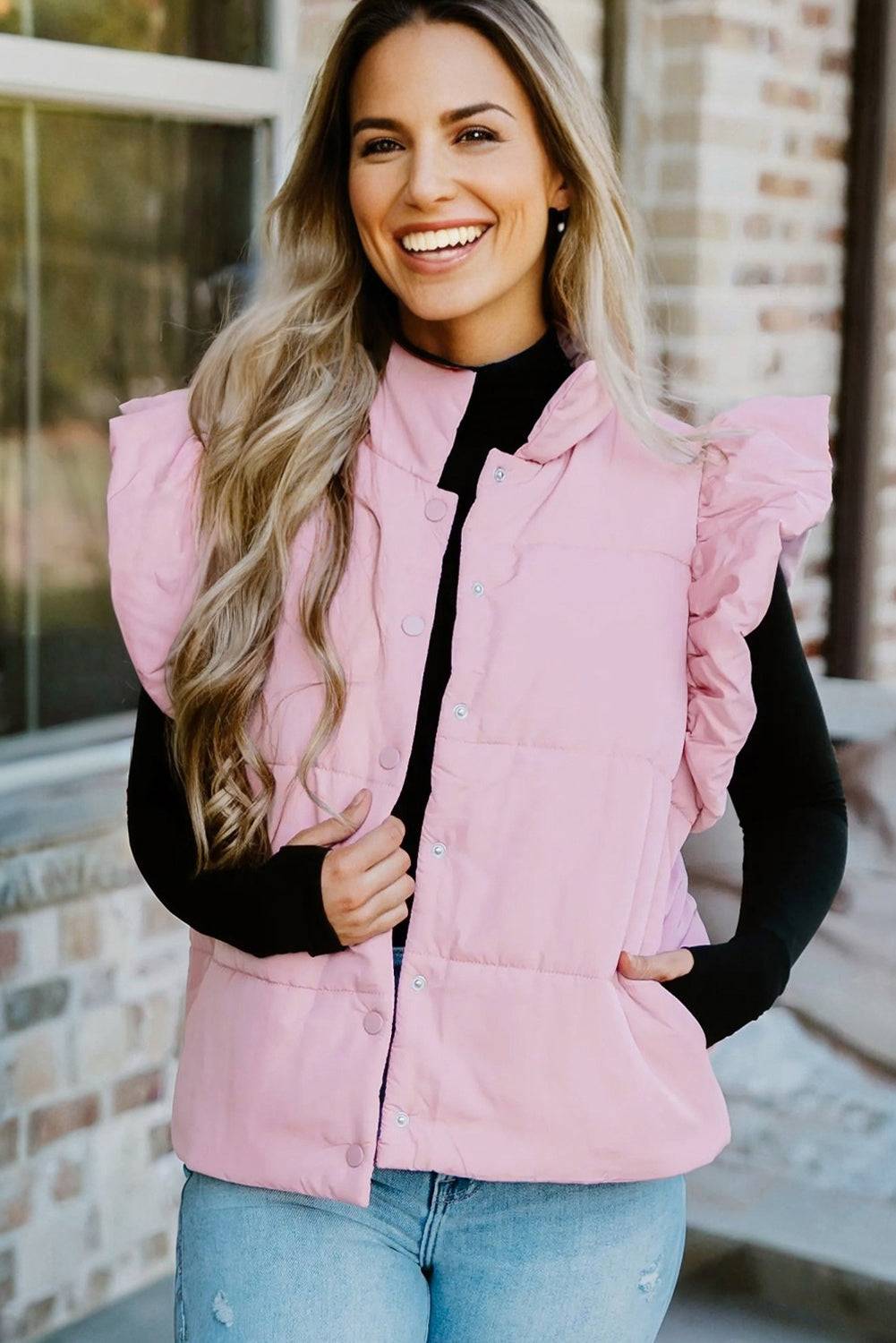 Woman wearing a pink puffer vest over a black top and jeans, standing in front of a brick wall.