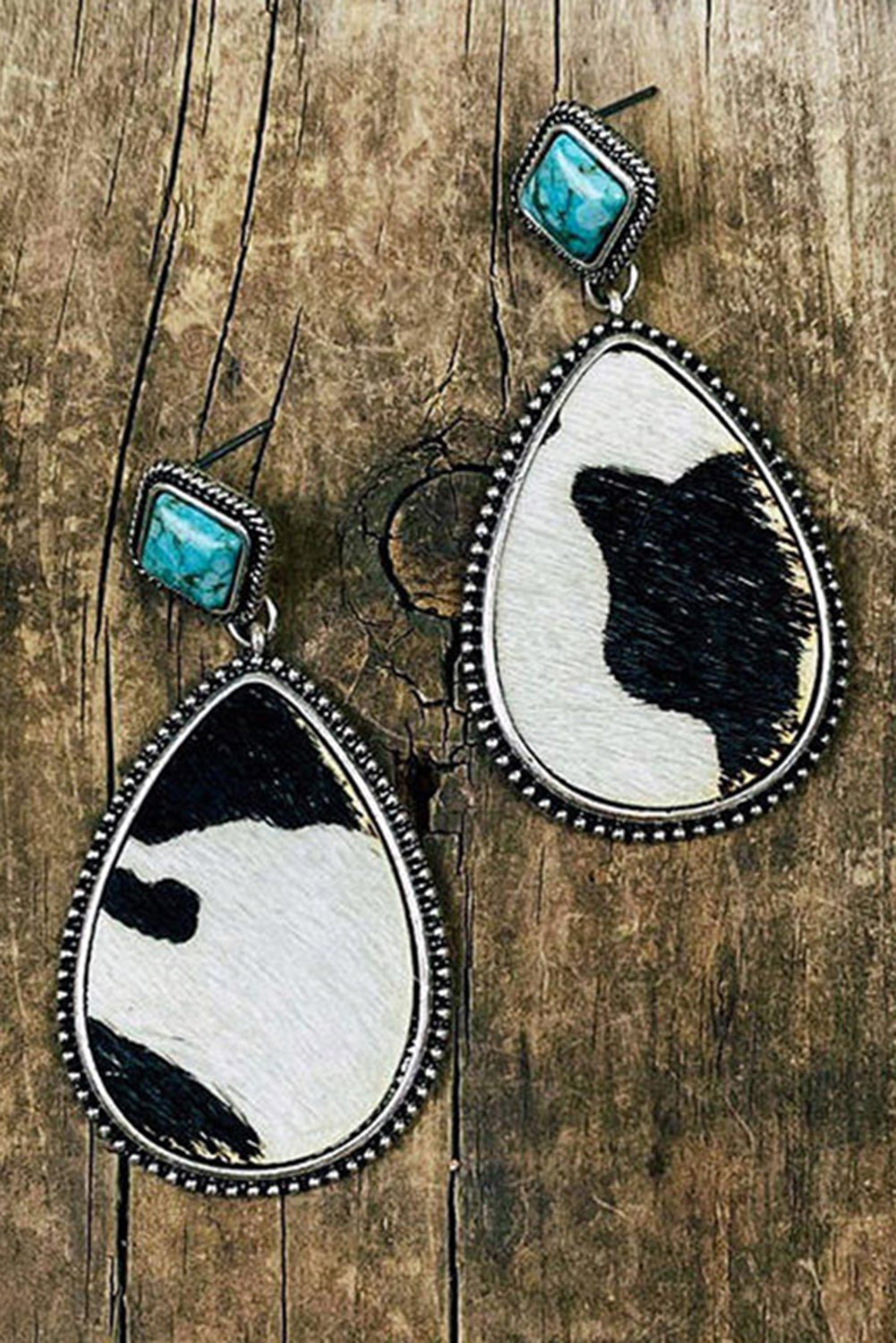 Teardrop-shaped earrings with black and white cowhide design and turquoise stones on a wooden background