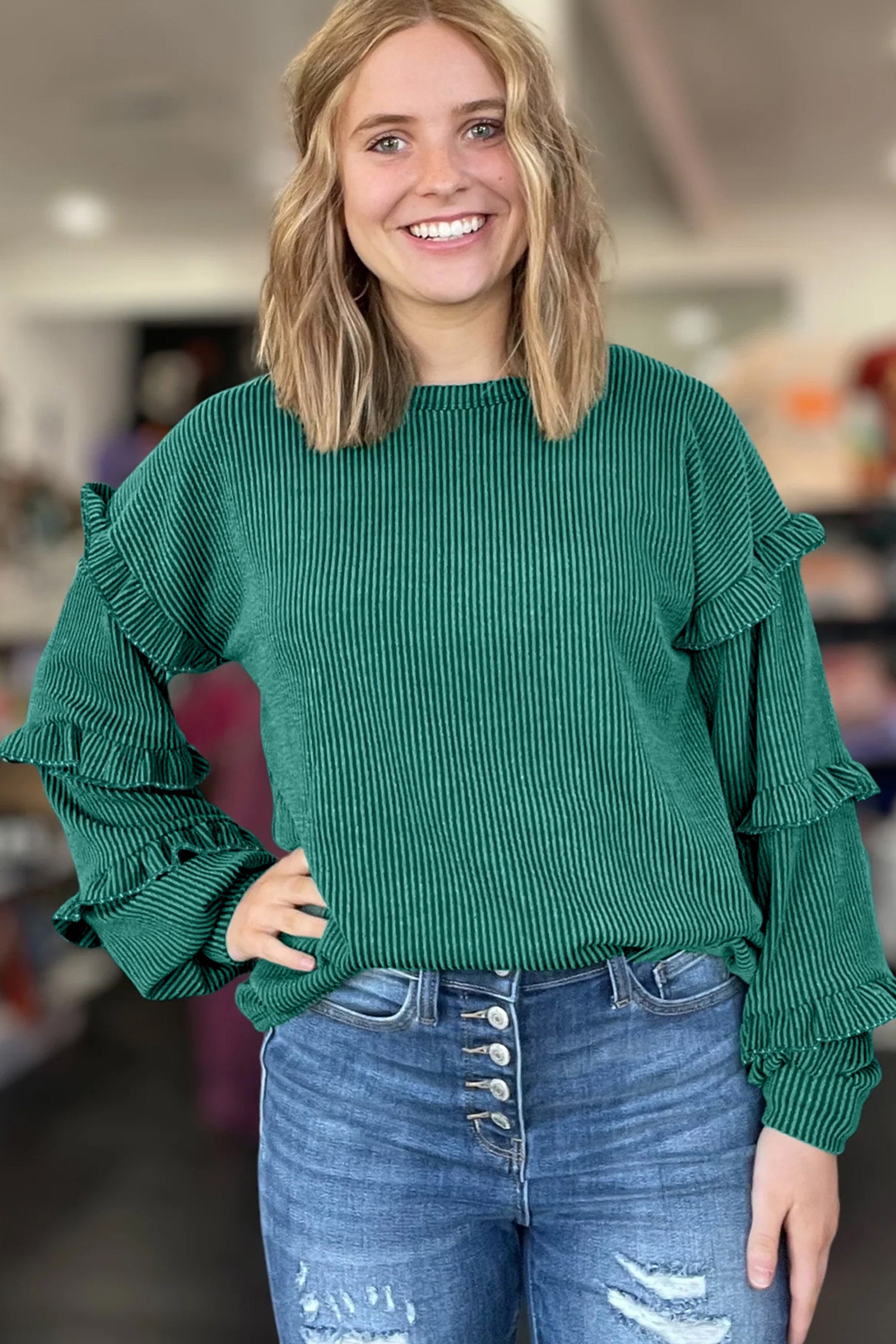 Woman wearing a green sweater with ruffled sleeves and blue jeans indoors.