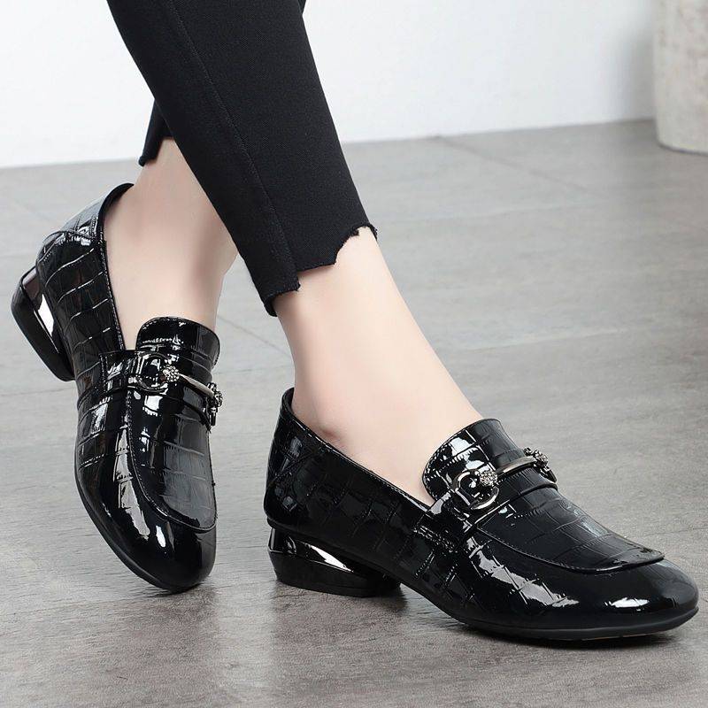 Black patent leather shoes worn with black pants on a light wooden floor.