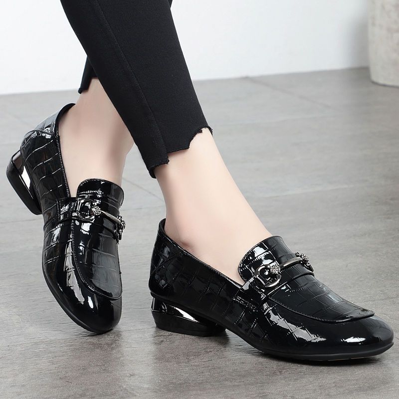 Black patent leather shoes worn with black pants on a light wooden floor.