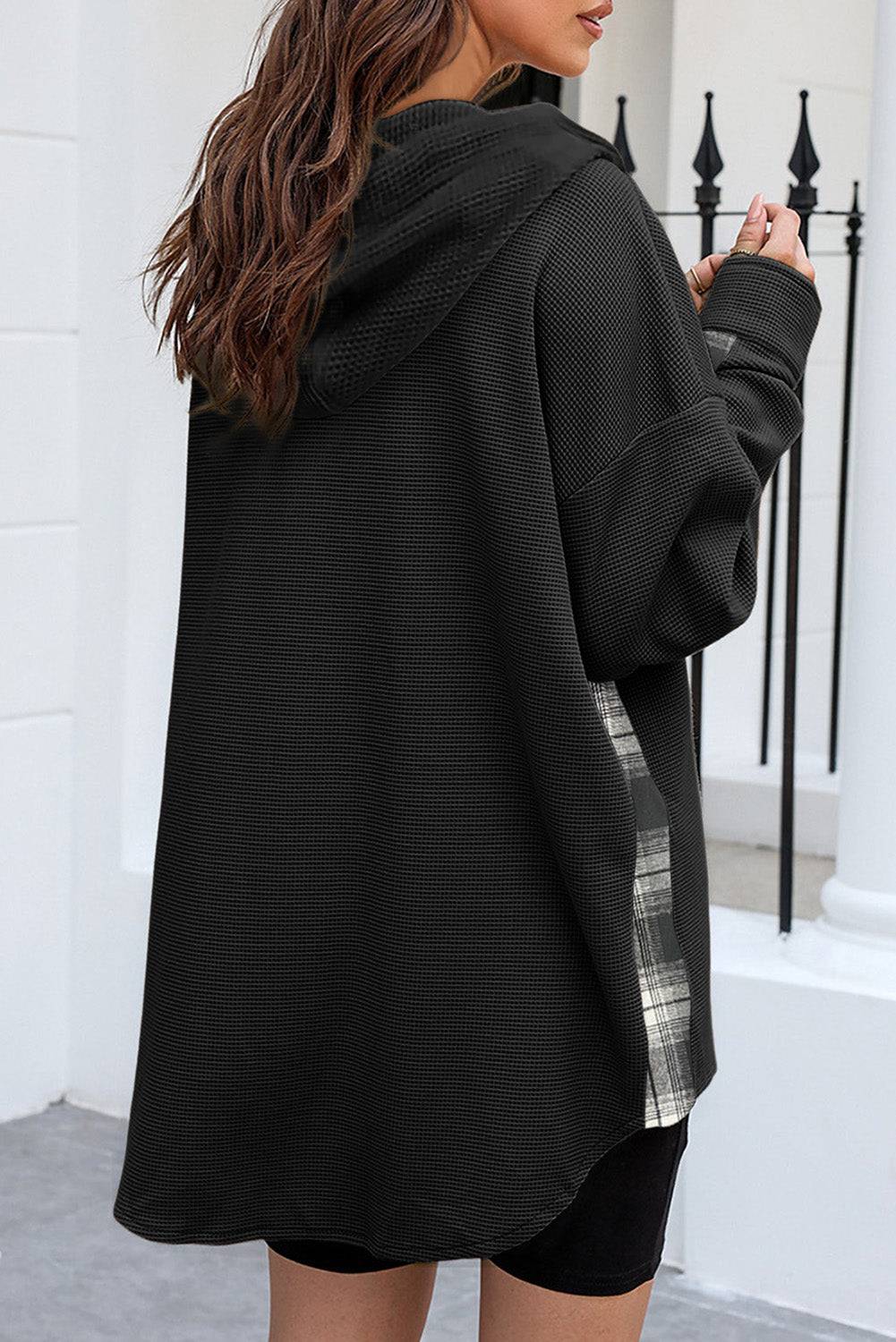 Person wearing a black oversized hoodie with plaid lining, standing on a white porch.