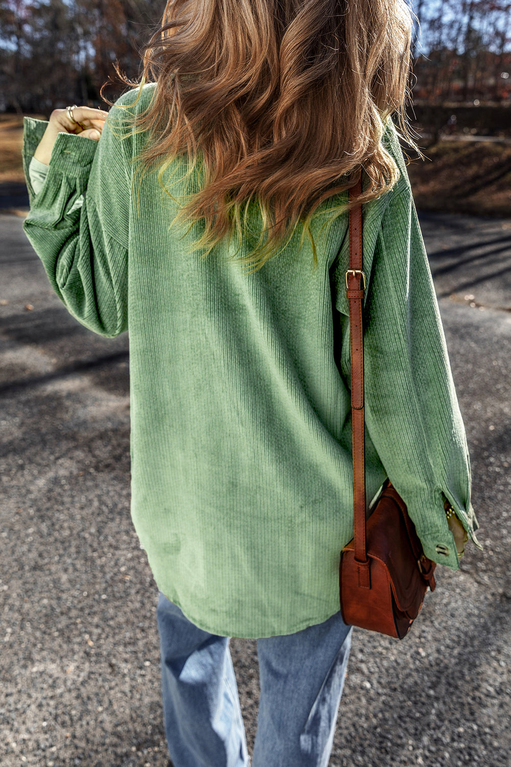 Person wearing a green corduroy jacket and carrying a brown leather bag on a road.