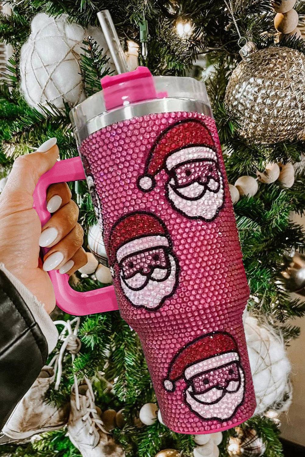 Pink blinged-out tumbler with Santa face designs held in front of a decorated Christmas tree.