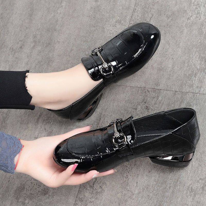 Black patent leather shoes with a decorative buckle held by a hand on a gray floor.