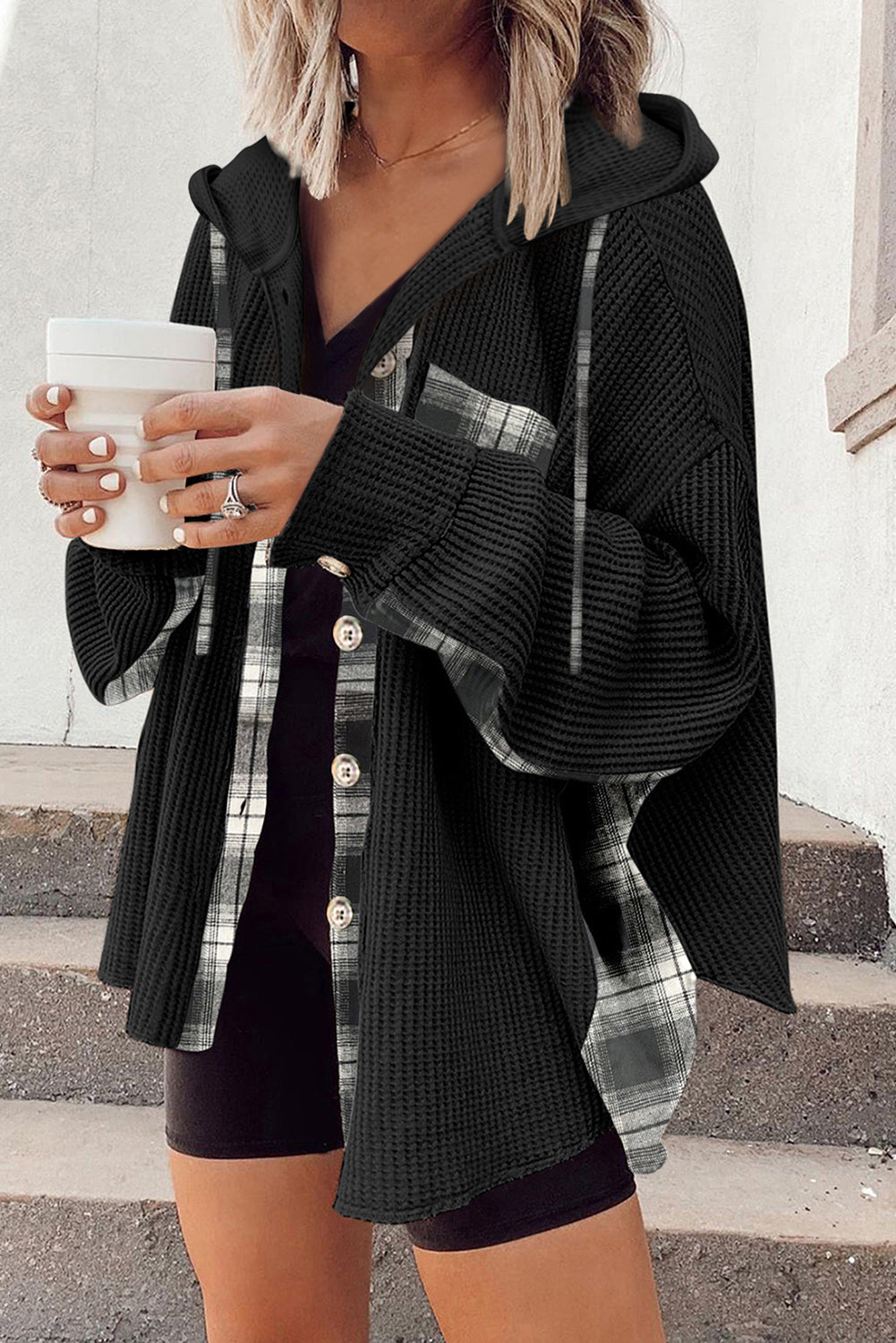 Person wearing a black cardigan over a plaid shirt, holding a white cup.